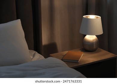 Stylish lamp and book on bedside table near bed indoors