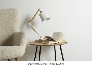 Stylish lamp with open book on wooden coffee table and soft armchair near white wall. Interior design