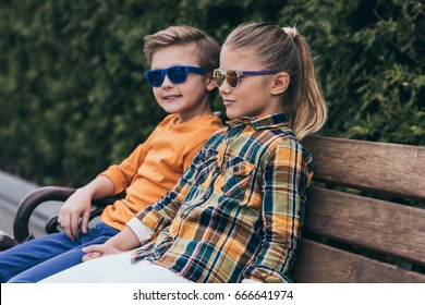 Stylish Little Kids In Sunglasses Sitting On Bench At Park