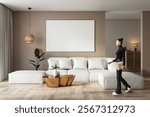 Stylish living room with white sofa, blank canvas on beige wall, wooden floor, decor accents, and a woman holding a notebook, modern interior