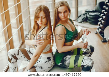 A stylish long-haired girl wearing a green sports suit sitting on a fitball near a large window in the gym with her beautiful girlfriend and they drinking a water