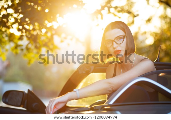 Stylish looking\
brunette woman with red lips wearing glasses, posing in cabriolet\
with a sun light. Empty\
space