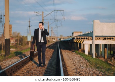 Stylish male businessman in a suit with a suitcase is walking along the tracks from the train and talking on the phone. Business walk and serious conversation. Late for the train.