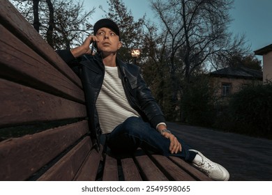 Stylish male model in leather jacket and sneakers posing on wooden bench at sunset. Urban lifestyle, or casual streetwear advertising. - Powered by Shutterstock