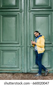 Stylish Man In Black Cap And Yellow Anorak Standing Sideways Against Green Background Holding Cell Phone Reading Funny Posts On His Social Network Page. People, Leisure, Technology, Style Concept