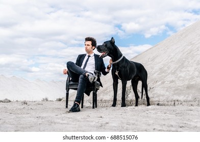 Stylish Man In A Black Suit Sits On A Chair In The Middle Of The Desert With A Big Dog. The Concept Of A Successful And Style