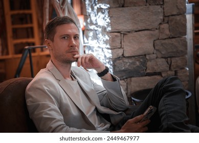 A stylish man in a blazer relaxes in a cozy room with a fireplace. He holds a smartphone, deep in thought. The setting exudes warmth and rustic charm. - Powered by Shutterstock