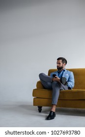 Stylish Man In Blue Trendy Suit Holding Smartphone And Sitting On Yellow Sofa