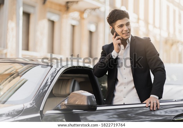 stylish man\
caucasian appearance businessman manager, sits in the car talking\
on the phone with a partner. Strict black suit, white shirt. Strict\
black suit, white\
shirt.