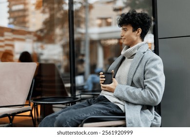 Stylish man with a cup of coffee sitting in a chair by a glass window looking out at the world - Powered by Shutterstock