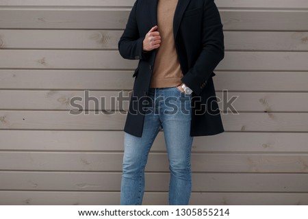 Stylish man in elegant fashionable black coat in a vintage knitted sweater in stylish jeans near a wooden wall. Spring collection of fashionable men's clothing. Close-up.