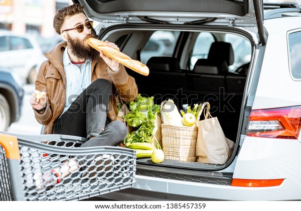 Stylish man\
enjoying food while sitting on the car trunk full of healthy\
products on the supermarket parking\
outdoors