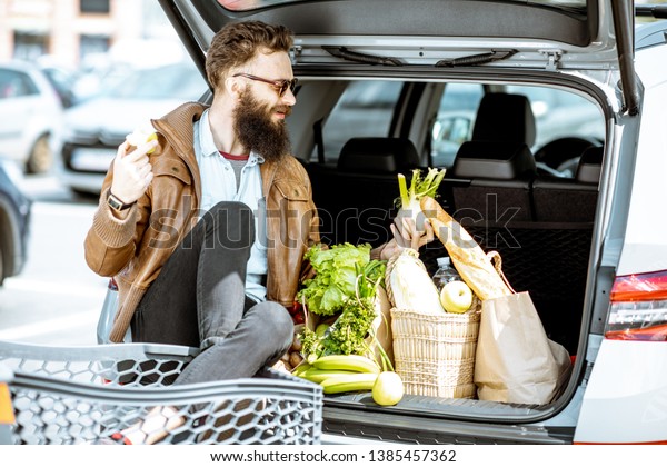 Stylish man\
enjoying food while sitting on the car trunk full of healthy\
products on the supermarket parking\
outdoors