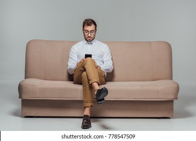 Stylish Man In Eyeglasses Sitting On Couch And Using Smarttphone Isolated On Grey