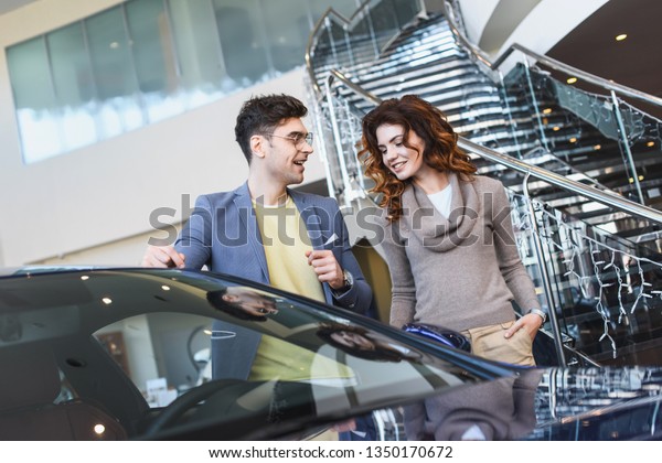 stylish man in\
glasses pointing with finger at car near curly attractive woman\
standing with hand in pocket\
