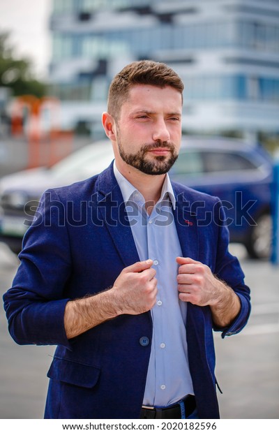 Stylish man in\
a jacket and blue jeans posing in an open parking lot. Car parking\
in the background. Business\
center
