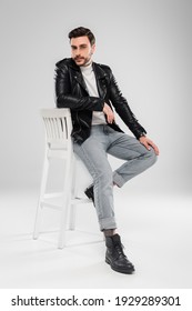 Stylish man in leather jacket looking at camera on chair on grey background 