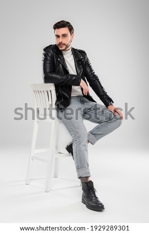 Stylish man in leather jacket looking at camera on chair on grey background 