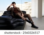 Stylish Man in a Modern Brown Suit Relaxing on a Black Sofa in a Minimalist Interior with Scenic Desert View Through Glass Windows.