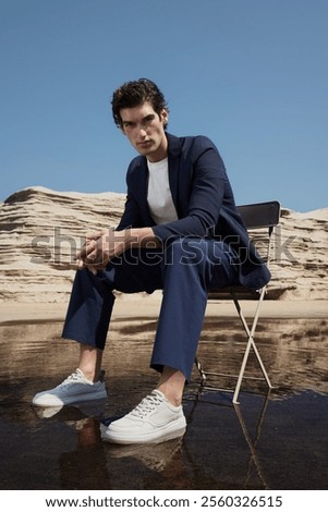Stylish Man in Modern Suit and Sneakers Posing Outdoors on Chair Against Rocky Landscape and Clear Blue Sky, Reflecting Contemporary Fashion.