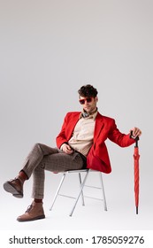 Stylish Man In Red Blazer With Crossed Legs, Posing With Umbrella While Sitting On Chair On Grey