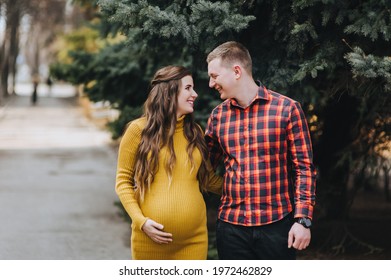 Stylish man in a red plaid shirt and a pregnant woman in a yellow dress stroll embracing on the street against a background of green pine trees. Pregnancy photography.