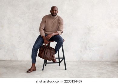Stylish man sitting on chair and looking camera. Confident fashion model in fashionable outfit turtleneck knitted sweater, plaid trousers, leather shoes and bag. Office clothes style for businessmen - Shutterstock ID 2230134489