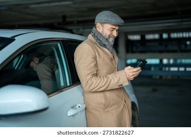 Stylish man smiling while operating a smartphone, standing beside his white car in a parking garage, embodying digital connection and urban convenience
