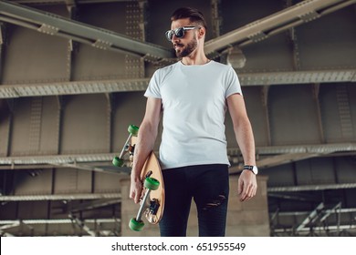 Stylish man in sunglasses and with a beard stands on the street with a long board. T-shirt mock up.