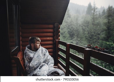 stylish man traveler in blanket relaxing on porch of wooden cabin in rainy day on background of woods in mountains. stylish hipster resting. space for text. atmospheric moment - Powered by Shutterstock