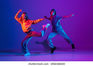 Stylish man and woman dancing hip-hop in casual sports youth clothes on gradient purple pink background at dance hall in neon light. Youth culture, hip-hop, movement, style and fashion, action.