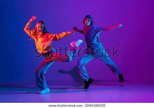 Stylish man and woman dancing hip-hop in casual\
sports youth clothes on gradient purple pink background at dance\
hall in neon light. Youth culture, hip-hop, movement, style and\
fashion, action.