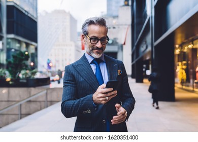 Stylish mature male in dark well-fitting business suit and glasses standing with slight smile on face in New York street and holding smartphone in hand  - Powered by Shutterstock