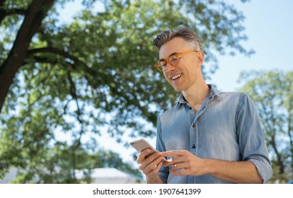 Stylish Mature Man Using Mobile App For Online Shopping, Chatting, Communication, Standing In Park. Happy Freelancer Receive Payment, Mobile Banking Concept  