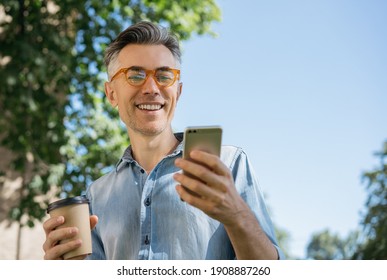 Stylish mature man using mobile phone, communication, reading news,   watching online movie, standing in park. Middle aged smiling hipster holding smartphone, drinking coffee outdoors - Powered by Shutterstock