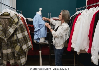 Stylish Mature White-haired European Woman Seamstress Trying On A Blue Blouse On A Mannequin In A Clothing Design Atelier, Standing Between Hangers With Clothes. Fashion Designer Business Start-up