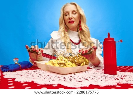 Stylish mature woman cutting with fork and knife fries and chicken against blue background. Fast food. Food pop art photography. Complementary colors. Concept of food, taste, creativity, retro style.