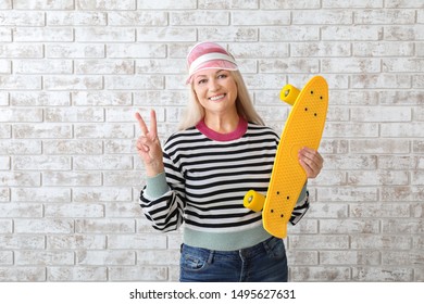 Stylish Mature Woman With Skateboard Showing Victory Gesture On Brick Background