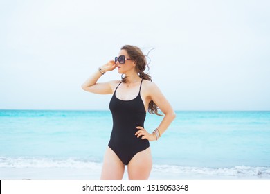 Stylish Middle Age Woman In Sunglasses And With Long Curly Hair In Elegant Black Bathing Suit On A White Beach Looking Into The Distance.