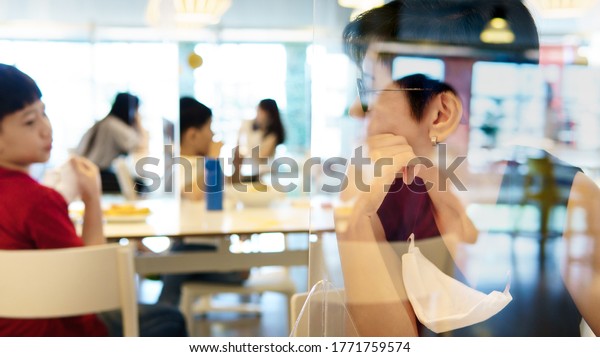 Stylish middle aged Asian mother talk to son who sit in\
separate table in food court with clear divider / barrier on table.\
New normal, Social & physical distancing during Covid-19\
pandemic 