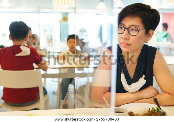 Stylish middle aged Asian woman with medical face mask\
sit separate from her kids in food court with clear acrylic divider\
/ barrier on table. New normal & Social distancing during\
Covid-19 pandemic 