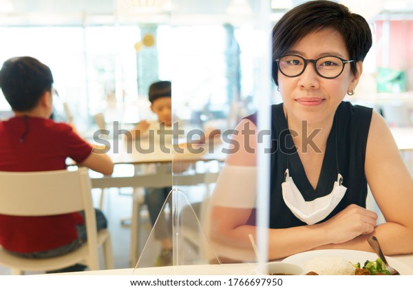 Stylish middle aged Asian woman with medical\
face mask sit separate from her kids in food court with clear\
acrylic divider / barrier on table. New normal & Social\
distancing during Covid-19\
pandemic