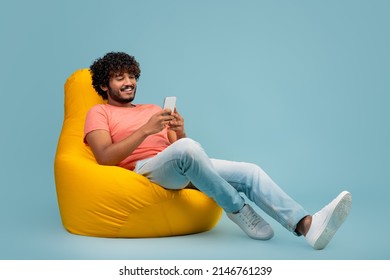 Stylish millennial hindu guy with curly hair sitting on yellow bean bag, using modern cell phone and smiling, using entertaining mobile application, blue studio background, panorama with copy space - Shutterstock ID 2146761239