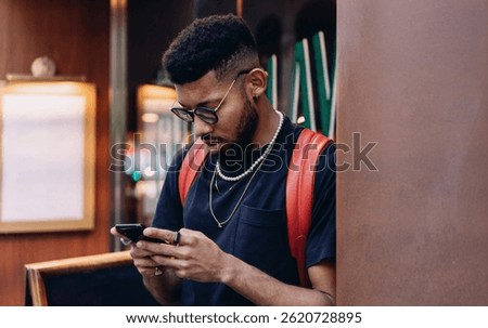 Stylish millennial male freelancer checking phone outdoors, wearing layered necklaces and rings, standing near café wall in city street. Modern tech and fashion blend naturally