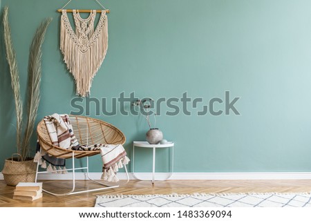 Stylish minimalistic interior of living room with design rattan armchair, palm leaf in basket, plaid, beige macrame on the wall, flowers and elegant accessories. Eucalyptus color of wall. Copy space. 