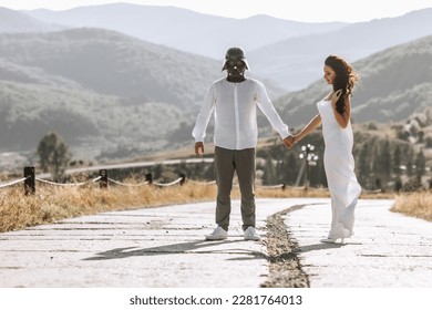 Stylish model couple in the mountains in summer. A man in a Darth Vader cap and a girl in a white silk dress against the background of a forest and mountain peaks.