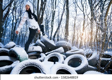 Stylish model posing on old used tires dumping in forest. Beautiful girl wearing in white warm hat and white blouse, jeans and sneakers on car tires covering snow.