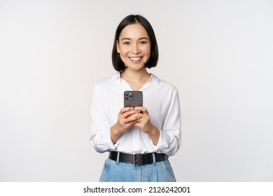 Stylish modern asian girl using mobile phone application, chatting on cellphone and smiling, standing in white blouse against studio background