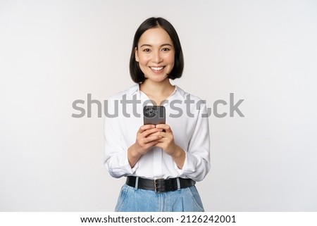Stylish modern asian girl using mobile phone application, chatting on cellphone and smiling, standing in white blouse against studio background