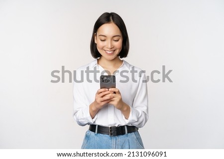 Stylish modern asian girl using mobile phone application, chatting on cellphone and smiling, standing in white blouse against studio background